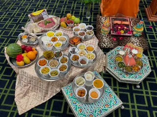 A photograph of a floor-level ritual food offering display on a navy and green patterned carpet. Many small white cups filled with different dishes sit on round silver trays covered by a pale gold cloth printed with Om symbols, including yellow sweets, orange curries, fried snacks, pickles, rice and sweets. Fresh fruit including watermelon, apples, oranges, mangoes and a dragon fruit sits on a silver tray at left. At upper right, a leopard-print cushion supports a red and gold tray with a pair of wooden sandals and rose petals, flanked by two decorated pots, and an orange cloth hangs behind. A silver octagonal tray with green and blue floral enamel holds steel bowls of vegetables and a pink-wrapped item. A small packet of pure dal or flour sits at top left. The arrangement reflects a Hindu naivedyam food offering.