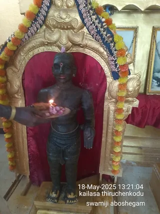 A photograph inside a temple shrine showing a dark stone statue of a standing male figure, bare-chested with a waist cloth and sacred thread, flanked by garlands of orange and yellow marigolds. A hand in a gold bangle holds a brass plate with a lit oil lamp and pink flower petals before the statue. The figure is set in a gold-framed arch with red velvet backing, with a gold-toned carved crown-like top and framed pictures on the wall behind. The statue is the central subject, with no living figure other than the offering hand. A timestamp and caption read 18-May-2025 13:21:04 and kailasa thiruchenkodu, swamiji aboshegam.