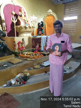 A man in a pink shirt and pink dhoti stands barefoot in a shrine room, holding up a dark book with a smiling woman's portrait on its cover. Behind him is a raised shrine with a golden lotus pedestal bearing a deity murti with a crown and garland, a trident, a black stone bull, small brass lamps, a kalasha, a bell and offering plates of flowers and fruit. A sign reads KAILASA SALEM BHARATH and a timestamp reads Nov. 17, 2024 5:35:02 p.m. Salem Tamil Nadu. No other person is shown.