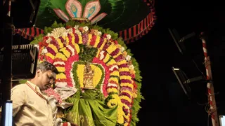 A photograph of a golden deity murti decorated with a dense frame of garlands in yellow, red, pink, purple and white flowers, with green leaves and a green silk cloth draped below. A green and pink umbrella-like canopy with red tassels hangs above. At the left edge a man in a cream-coloured dhoti and shirt with a red sacred thread stands with eyes lowered, holding a silver vessel with pink flowers. Floodlights and a red-and-white striped pole are visible at the right, and the background is dark.