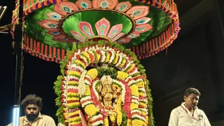 Night photograph of a golden deity murti of Ganesha, the elephant-headed god, framed by a dense arch of marigold and mixed flower garlands in yellow, white, red and pink, with green leaves. A large green fan-shaped canopy with peacock-style painted eyes and pink petals, edged with red and purple tassels, hangs above. Two men stand at either side: on the left a bearded man with curly hair in a white shirt, on the right a man in a white shirt and white dhoti-style cloth wearing a thin sacred thread, both looking downward in devotion during ritual service. The background is dark.