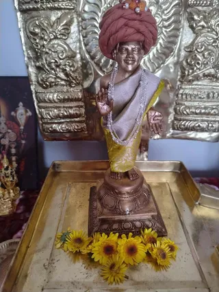 A bronze-toned statue of a standing male figure wearing a rust-red turban decorated with rudraksha beads and pearls, a grey shawl over a yellow-gold brocade garment, and layered pearl and silver chain necklaces. The statue raises one hand in blessing and holds the other palm open at its side, standing on a lotus-style pedestal on a brass tray. Yellow chrysanthemum-style flowers are scattered at its base. Behind it is an ornate embossed brass backdrop, and to the left a framed image of a deity with a gold-decorated figure. Statue, not a physical person.
