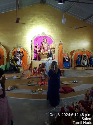 Interior of a temple hall with gold-painted walls and a corrugated metal roof, with ceiling fans. A central shrine niche with a pink backdrop holds a seated deity murti decorated with garlands and a trident, with a placard reading "KAILASA SALEM BHARATH" beside it. Flanking niches hold dark-stone and painted deity murtis with garlands, including figures in blue and white. A woman in a navy blue sari with flowers in her hair stands in prayer with her back to the camera before the steps of the shrine, where brass lamps, plates and offerings sit. At the lower right, a display of painted clay dolls and figurines is arranged on a red cloth. A timestamp reads Oct. 6, 2024 6:12:48 p.m., Salem, Tamil Nadu. Two other partially visible people with folded hands stand at the left edge.