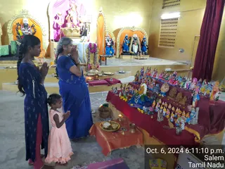 A photograph inside a temple hall with yellow-gold walls and arched niches holding deity murtis, one above shows a large figure with a pink-and-white draped backdrop. Three people stand with hands folded in prayer: an adult woman in a blue saree and a girl in a dark blue kurta with a long side braid, and a small child in a pink dress. To the right, a long table draped in maroon and gold cloth displays a Golu arrangement of many painted clay dolls and figurines, with a blue-robed figure at center. Brass plates, oil lamps and a kalasha sit on the floor and steps, indicating puja. A printed timestamp reads Oct. 6, 2024 6:11:10 p.m. Salem Tamil Nadu.
