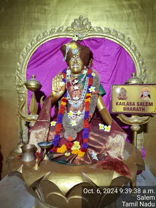 A golden statue of a seated figure is the main subject, placed on a gilded lotus pedestal in front of a golden arch and magenta cloth backdrop. The figure wears a brown headdress with a jewelled ornament, a maroon and gold brocade garment with a green blouse, layered white and multicoloured bead necklaces, and a long rudraksha-like cord; it raises one hand in a blessing gesture. Marigold and purple garlands and white plumeria flowers adorn the statue. A brass kalasha, a small brass lamp and a brass bell sit on the pedestal, and a gold lamp stand flanks the base. A plaque on the right shows a smiling saffron-turbaned figure's portrait beside the caption KAILASA SALEM BHARATH. The statue is a depiction, so no physical people are present. A timestamp reads Oct. 6, 2024 9:39:38 a.m., Salem, Tamil Nadu.