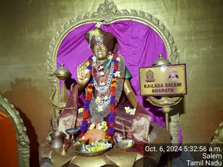 A golden statue of a seated figure in a brown turban with a jeweled ornament and a central red mark, raising its right hand in blessing. It wears layered garlands of red, orange, purple and white flowers, a gold necklace, and a brown shawl over a green blouse, seated before a magenta drape within an ornate gold arch. In front stand a brass pot, oil lamps, a brass plate of marigolds and white plumeria, a lit candle, and an orange offering. A rectangular placard reads KAILASA SALEM BHARATH beside a small printed portrait of a smiling figure in saffron. A timestamp in the corner reads Oct. 6, 2024 5:32:56 a.m. Salem Tamil Nadu. No physical people are visible.