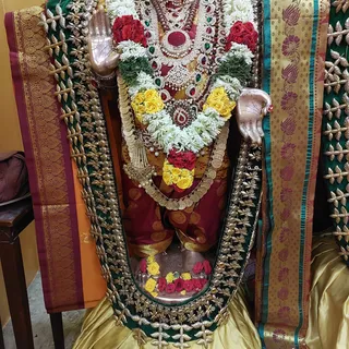 A photograph of a standing metal deity murti. The murti wears layered gold and jewelled necklaces set with red and green stones, a long pearl-and-gold ornament, and a thick garland of white jasmine, red roses and yellow roses. Its raised hands are visible on either side, and rose petals are arranged at its feet on a silver base. A gold-and-maroon silk cloth and a yellow satin backdrop frame the figure against a yellow wall.