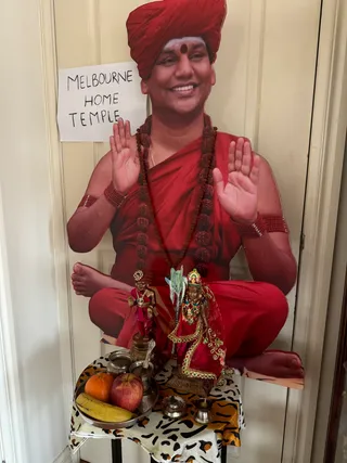 A photograph of a life-size cutout standing against a cream panelled door, showing a smiling man in a red turban with a red bindi, red robes and a long rudraksha mala, raising both palms in blessing. A handwritten paper sign is attached to the door beside the cutout. In front of it stands a small table draped in a leopard-print cloth holding a silver tray with an orange, an apple and a banana, a small brass kalasha-style vessel with spoons, and two brass oil lamps. Two metal deity murtis dressed in red and gold stand on the table, one holding a staff-like object and a flower. The cutout depicts a depiction of Swamiji (probable) from its red turban, rudraksha malas and saffron-red robes, shown on a printed panel rather than as a physical person.