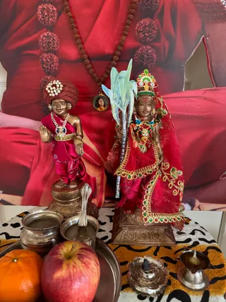 A small household altar photographed on a leopard-print cloth. Two bronze-finished murti figures stand side by side: on the left a child-like figure in a red turban with pearl ornament, red satin garment, beaded necklaces and a raised hand; on the right a deity murti in a red sari-like drape with gold embroidery, a jeweled crown and a green-and-red beaded garland, holding a tall green stalk. Behind them a large printed image shows a seated figure in red robes with a rudraksha mala, and a small locket pendant hangs from the mala. In front are a silver pot with spoon, a silver bowl, a red apple, an orange, a silver flower-shaped incense holder and an oil lamp. No people are visible.