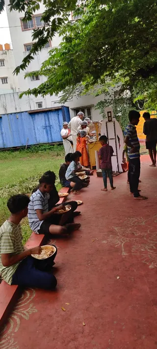 Outdoor photograph of an Anna Daan food distribution on a red-painted courtyard path beneath a large tree. Several boys sit along a low red ledge eating from metal bowls, one in a green-and-white striped shirt in the foreground. Behind them, volunteers in white aprons and hair nets serve food from a yellow counter, with a woman in an orange sari among them. A blue shipping container and a white multi-storey building stand in the background, and more boys stand at right. Patterned floral motifs are painted on the ground.