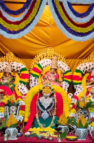 A photograph of a decorated deity alankaram for Navaratri inside a hall with a gold cloth canopy. Three goddess murtis sit on golden thrones framed by garlands of yellow, red, green and white flowers with dotted white blossoms. The central murti wears a silver-and-stone ornamented bodice, a jewelled crown, a long bead necklace and a yellow face; a smaller seated figure in a pink sari sits above. Kalashas wrapped in mango leaves and yellow flowers flank the altar, with brass oil lamps, a bell, a brass vessel and small offering bowls with yellow petals on a red cloth. No people are visible.