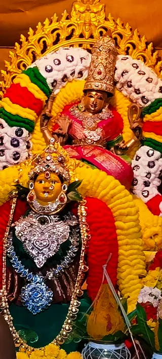 A photograph of a decorated deity alankaram at a temple shrine. Two golden murti figures are adorned with dense yellow, red and green marigold and flower garlands and white flower garlands studded with dark beads. The upper murti wears a gold crown, a pink and red silk sari, and layered gold necklaces, with gold arms raised. The lower figure wears a jeweled silver-and-stone breastplate, a blue gemstone pendant, a jeweled crown and red and green earrings, against a dark green cloth. A golden arched prabhavali frames the figures, and a yellow kalasha with a white thread stands in front. No people are visible.