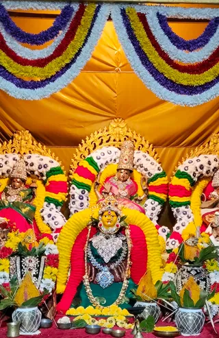 A photograph of a decorated deity alankaram at a temple shrine. A yellow-faced central deity murti sits under a yellow and red floral arch, wearing a green sari, a silver heart-shaped chest ornament, layered silver necklaces and a jewelled crown. Behind it, a golden backdrop hangs in pleats, with strings of white, yellow, red, green and purple flower garlands draped above. Two smaller murtis flank the central figure, each in pink, red or green garments with crowns. Brass and metal pots decorated with mango leaves and marigolds stand at the base, with small oil lamps, a brass vessel, flower petals and a plate of offerings on the red cloth. No people are visible.