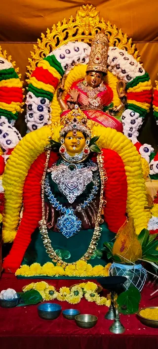A decorated deity murti set on a red-draped altar in a shrine. The central figure is a golden-faced goddess with a jewelled crown, a large silver-and-blue crystal necklace and a gold bead garland, seated within a thick arch of yellow and red marigold-style garlands studded with white and sparkling beads. Above and behind, a second smaller golden figure in a pink and red sari wears a tall jewelled crown and holds objects in raised hands. Yellow marigold petals lie before the murti, with brass bowls, a brass oil lamp, green leaves and a decorated kalasha with mango leaves at the right. No physical people are visible.