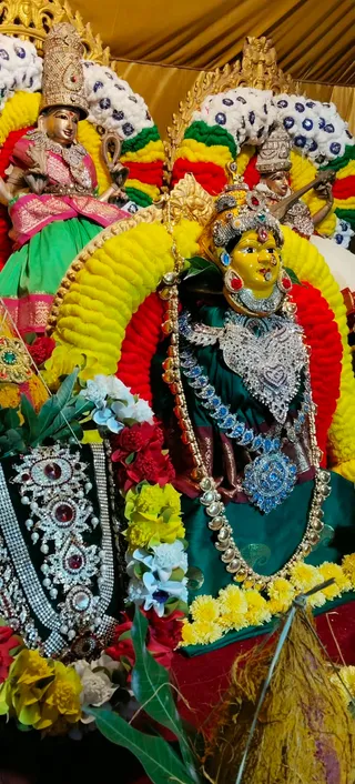 Close photograph of festive deity alankaram at a KAILASA Hyderabad Navaratri setting. Central deity murti with a yellow face, jeweled crown and heavy silver-toned and blue-stone necklaces, dressed in a dark green silk garment. It is framed by a large arch of yellow, red and green marigold garlands. Flanking figures wear pink and green silk with silver ornaments; one holds a veena. Decorative silver and gemstone jewellery, white flowers and mango leaves surround the lower left, and yellow marigold garlands drape the base. No physical people are visible.