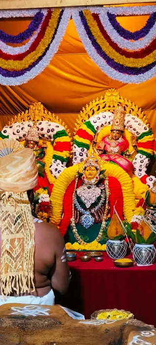 A photograph of a decorated deity alankaram at a temple shrine. In the centre, a golden-faced deity murti wears a heavy silver breastplate, gold necklaces and a jeweled crown, framed by a yellow and red flower arch. Behind her, a second murti in a pink sari with a tall gold crown is seated on a golden throne with floral garlands in red, yellow, purple and white. Orange and gold draped fabric hangs overhead. On the red table in front are silver pots with mango-leaf and marigold decorations, brass bowls, a plate of turmeric or kumkum and a basket of yellow flowers. At the lower left, a man with his back to the camera wears a cream and gold shawl over bare skin and a white dhoti, standing before the altar.