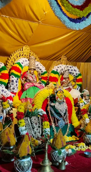 A decorated shrine under a golden canopy with yellow, red and white flower garlands arching behind the figures. Three devi murti sit on thrones: the central one wears a green and dark-green silk sari with heavy silver jewellery and a blue pendant, a crown and a yellow face; a figure on the left holds a lotus and staff-like object in a pink and green sari; another on the right holds a veena in a white garment. Kalasha pots wrapped in yellow cloth stand at the base beside a brass oil lamp, marigold garlands, and a red cloth covering the floor. No people are visible.