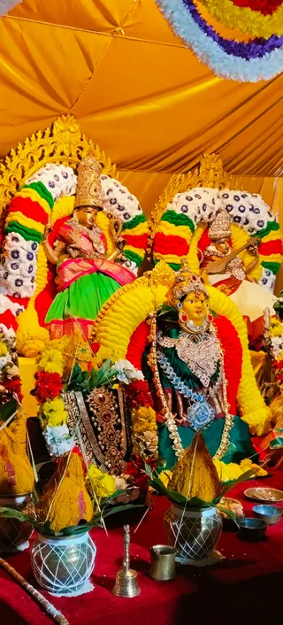 Photograph of a festive altar decorated for Navaratri under a yellow canopy. Three deity murtis are dressed and adorned: the central figure wears a green-and-dark-green sari, a silver chest ornament, a blue stone necklace and a red-jeweled crown, flanked by a murti in a green skirt and pink-red sash and another holding a veena in red clothing. Each is framed by an arch of yellow, red, green and white flower garlands on gilded thrones. In front are brass kalasha pots topped with yellow-dyed coconut and mango leaves, a brass bell, small brass cups and a red cloth covering the platform. The scene reflects a devi alankaram.