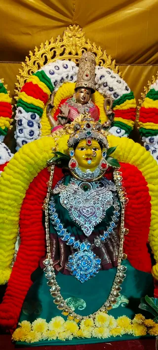 A photograph of a goddess murti decorated for Navaratri alankaram. A yellow-faced deity figure wears a crown studded with red and green stones, a jeweled silver-and-blue necklace, and a dark green silk drape with gold trim. Behind her, a standing golden-skinned figure in a pink sari holds a lotus-like object and a pot, with a golden arch behind. The murti sits within an arch of yellow, red, green and white flower garlands, with a row of yellow chrysanthemums at the base and green leaves at the face. No people are visible.