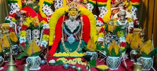 A decorated deity murti is the central focus of a devi alankaram arrangement. The central figure has a yellow face, a silver crown, and a silver heart-shaped chest ornament over a green garment, with layered silver necklaces and a blue gem pendant. A yellow and red garland frames an arch behind her. Flanking figures wear red, white and green floral garlands and silver ornaments. In the foreground are brass oil lamps, bronze bowls, a brass kalasha-style vessel, marigold and yellow flowers, and green leaves arranged in decorated pots. No people are visible.