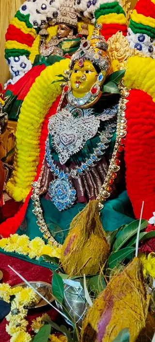 A decorated deity murti of a goddess, seated and framed by arches of marigold and bead garlands in yellow, red, green and white. The deity wears a jeweled crown, a silver-and-blue embellished chest ornament, a green and maroon garment and a gold-beaded necklace. Two coconuts with fibrous husks sit in front, surrounded by yellow marigold flowers, mango leaves and a silver plate. A second smaller figure appears in the background at top. No people are visible.