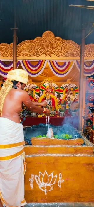 A bare-chested male priest in a white dhoti with gold borders and a yellow head cloth stands at left, pouring liquid from a vessel onto a fire pit. The homa altar is a tiered yellow-ochre mud platform with a white lotus drawing and kumkum dots, with green leaves placed on top and smoke rising. Behind it, three decorated deity murtis sit under a gold carved arch draped with garlands in red, yellow, green and purple. Several kalashas and brass vessels stand before the deities. The priest wears a rudraksha-style mala and red-and-gold wrist threads. No a person candidate is visible.