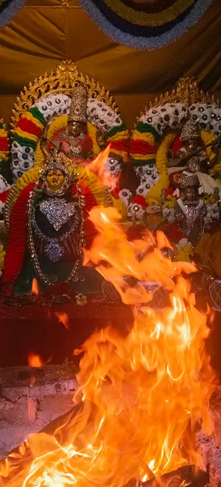A large fire pit in the foreground blazes with tall orange flames before a decorated shrine of deity murti adorned with silver and jewelled ornaments, crowns, and garlands of red, yellow and green flowers with white beaded arches. A gold canopy with a blue and yellow floral border hangs above. The central figure is a dark-faced goddess murti with a silver heart-shaped breastplate and a crown, flanked by additional murtis. The scene suggests a homa fire ceremony, with no people visible.
