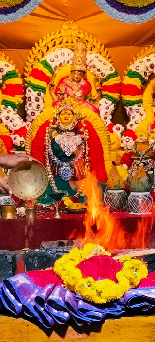 A photograph of a decorated deity murti of a goddess seated within a shrine framed by yellow and red marigold garlands and white floral arches. The murti wears a silver and jewelled ornamental crown, a dark green sari with silver necklaces, and a large embellished breastplate. Above it stands a smaller golden figure in a pink garment. In front, a sacred fire burns in a fire pit, and a hand at the left holds a brass vessel pouring offerings. Yellow marigold garlands wreath a red cloth bundle beside the fire, and blue and pink silk cloth lies on the ground. Silver-patterned kalasha pots stand on the right, with a brass oil lamp below the murti. The scene reflects a homa ritual during Navaratri devi alankaram.