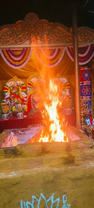 A large fire blazes in a clay-walled homa pit in front of a decorated shrine at night. Behind the flames sits a deity murti adorned with garlands of white and red flowers, and a golden canopy with draped flower garlands frames the altar. A carved golden arch tops the shrine, and a painted panel with floral and elephant motifs stands at the right. No people are visible in the frame, and the fire is the central subject of this chandi homa scene.