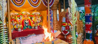 A photograph of a homa fire burning in a brick fire pit in front of a decorated shrine. On the left, several deity murtis in red and green garments with gold crowns and floral garlands sit on a red-draped platform, flanked by kalasha pots and marigold garlands. Behind the fire, a golden ornate throne holds a figure in a red turban with a multicoloured floral garland, and palm fronds frame the scene. Colourful striped fabric hangs from a golden pillar on the right, and an orange cloth backdrop covers the top. No people are clearly visible standing in the scene apart from the seated figure on the throne.