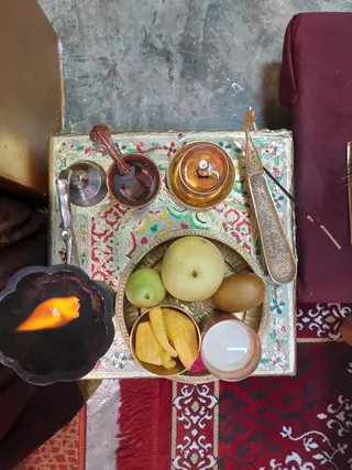 Overhead photo of a puja setup on a gold-and-red patterned square tray with floral motifs. A brass plate holds whole pears, a kiwi-like fruit, sliced yellow fruit, and a small white-filled cup. Nearby sit a silver bell, a copper cup with a small ladle, a brass oil lamp with a flame, and an ornate brass spoon, with incense sticks at right. A dark ceramic lamp with a bright flame sits at left on a red rug. No people are visible.