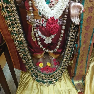 Close view of a deity murti dressed in a deep maroon silk cloth with gold-embroidered borders, adorned with a large white jasmine garland, a cluster of red roses at the chest, a silver beaded mala, a jewelled crescent-shaped ornament and a pearl-and-diamond pendant. The lower part shows yellow and red roses scattered at the base in a silver vessel-like setting. A gold-and-green embroidered sash frames the figure, and a metallic hand with an outstretched palm appears at the right beside a teal and gold patterned cloth. No people are visible.