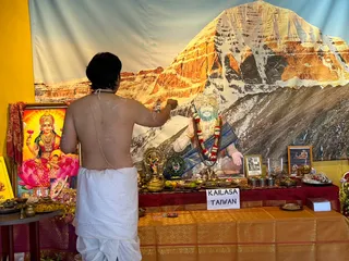 A bare-chested man in a white dhoti with a sacred thread stands with his back to the camera at a home altar in front of a large mountain backdrop. He is raising one arm, apparently marking or sprinkling near the altar. On the altar are a framed goddess image with a lotus seat and red-gold background, a framed portrait of a bearded sage wearing a red-and-white flower garland, small brass lamps, a brass kalasha, fruit, bowls, flower offerings and a sign reading KAILASA TAIWAN. A small framed black-and-white photo sits beside the sage portrait. The altar is covered in red, gold and maroon cloths.