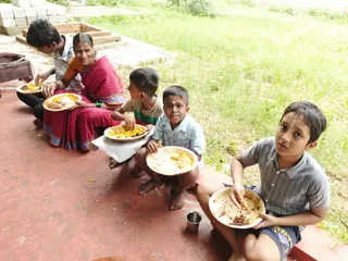 A photograph of an outdoor Annadan meal on a red-painted concrete ledge beside grass and a brick step. Five people sit eating from disposable leaf-style plates: a woman in a magenta sari with an orange border and green bangles, and four children, including a boy with a white forehead mark on the right. Plates hold rice with yellow and brown dishes. A metal drum stands at the left edge, and a small steel cup sits on the ground in front.