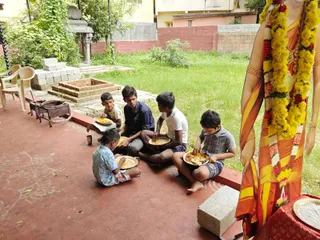 Four boys and one smaller child sit on a red-tiled courtyard floor eating from metal plates of food. Their clothes include a blue shirt, white t-shirt, checked shirt and light blue patterned shirt. Beside them stands a large life-size image in orange and yellow sari with a yellow marigold garland and red roses, and a metal plate rests on a red cloth. Behind them are green grass, a brick fire pit, a small stove, plastic chairs and a pillared stone shrine structure by a pink wall.
