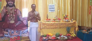 A photograph of a man in a white dhoti and shawl with bare upper body, standing with palms pressed together in namaskar in front of an altar draped with a yellow embroidered cloth. The altar holds small figures of deities, flower decorations, brass plates and bowls of fruit and food offerings, including pineapple, mango, watermelon and bananas. Behind him is a large printed backdrop of a seated saffron-robed figure with a red turban, rudraksha and gold garlands, resting on a tiger-skin seat. A paper sign on the yellow curtain reads KAILASA TRINIDAD & TOBAGO. The scene suggests a puja offering; one person is visible.
