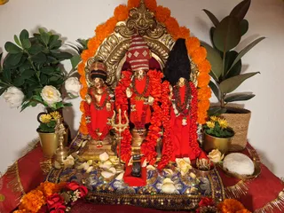 A photograph of a devotional shrine arrangement with three deity murtis in red garments, adorned with red and rudraksha-style beaded garlands, set before an arched golden frame edged with marigold garlands. The central murti holds a trident-like object in front of it; a small brass bell stands at left, and white roses and rose petals are scattered on a blue embroidered cloth. Potted plants, yellow chrysanthemums, a white offering on a brass plate, and red flowers frame the altar. No people are visible.