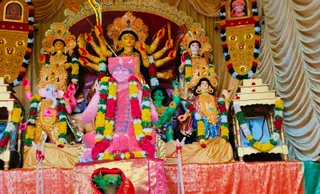 A photograph of a Durga Puja altar at KAILASA Hyderabad, dominated by a large multi-armed Durga murti with a golden crown and weapons, flanked by Ganesha with an elephant head, a green-skinned figure, and two other deity murtis. Central in front is a seated figure in a pink-red shawl and rudraksha mala, with a red-and-white tilak marking, the figure's clothing and garlands in red, yellow, and white flowers, which is a statue or figure rather than a clearly visible living person. Framed portraits hang on the sides, one showing a figure in orange with a turban, and the altar is decorated with gold-trimmed panels, golden drapes, marigold and rose garlands, and a red-and-gold cloth on the platform.