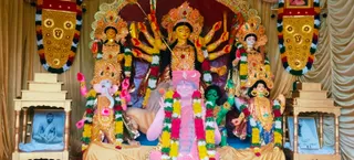 A decorated Durga Puja pandal shrine with a large multi-armed Durga murti in red and gold at centre, holding weapons, with Ganesha in pink and gold to the left and figures in bright costumes flanking her. Garlands of yellow, red and white flowers hang across the figures, and gold-framed banners with ornate motifs decorate the upper corners beside gold draped fabric. A pink standing figure in front wears a crescent-topped headpiece with a garland and is partly obscured by a shoulder-height arrangement. Two framed black-and-white photographs stand on small gold tiered pedestals on either side, one showing a seated man in meditation posture. No physical people are visible in the scene.