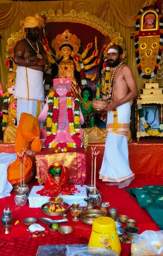 Photograph of a Navaratri Durga puja set up with a golden-framed deity murti of Durga, multi-armed and crowned, behind a pink-painted printed portrait of a smiling figure in a saffron turban with garlands on a golden seat. Two men in white dhotis stand on a red-draped stage: the left figure, bare-chested with a yellow turban, rudraksha-style beads and a cloth tied at the waist, holds a sword; the right figure, bare-chested with a white forehead marking and garlands, holds a copper vessel. A kneeling person in an orange cloth sits at the front. Brass bowls, flowers, lamps, a coconut, a yellow pot and a framed photo with garlands surround the altar. Three people are physically present.