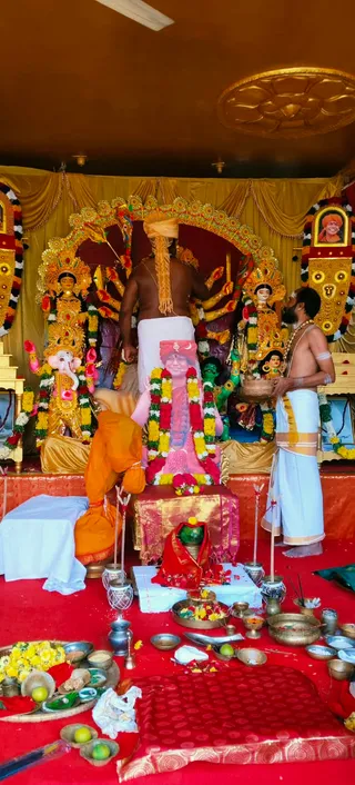 A photograph of a Durga Puja setup in a decorated shrine. A bare-chested man in a white dhoti with a golden-yellow turban-like wrap stands at the centre, facing a framed portrait of a figure in orange and red garland-draped clothing set on a pink-draped stand. A second man in a white dhoti with a gold border, bare-chested and holding a brass vessel, stands at right. A person in an orange robe bends at left. Golden deity murtis with multiple arms, including a Ganesha figure at left and goddess figures with red and gold garlands, stand behind. Red floor cloths hold brass bowls, flowers, lemons, betel leaves and oil lamps. A framed portrait with an orange-turbaned figure appears at upper right.
