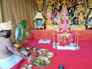 A seated priest bare-chested with a white dhoti and a pale cloth turban performs a puja before an altar draped in red cloth. Behind the altar stands a large deity murti of Durga with multiple arms holding weapons, flanked by figures including Ganesha with a yellow garland and other goddess figures decorated with marigold garlands. Brass plates and bowls hold offerings, leaves, kumkum and turmeric, and lit incense sticks rise from small holders. A small framed portrait of a man in a seated pose sits on a wooden stand to the left of the deities. A potted plant stands at the left edge. Golden drapery hangs in the background.