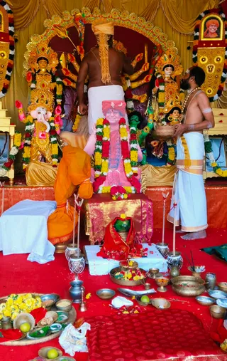 Photo of a Navaratri Durga Puja worship set-up. A shirtless priest in a white dhoti with a golden shoulder cloth stands at a golden shrine. Behind him, a multi-armed golden Durga murti and a golden Ganesha murti flank the altar, and a green-faced figure stands to the right. Strings of yellow and red marigold garlands drape the shrine. In the centre, a large framed printed portrait of a smiling man in saffron turban, rudraksha-like beads and ornate garland sits on a gold-trimmed cloth; the portrait is a depiction, not a physical person. A second priest in a white dhoti with a gold border, bare-chested, holds a basket of flowers on the right. A kneeling figure in orange garments bends at the left. Brass bowls with flowers, fruit, kumkum and offerings, incense sticks, and kalasha-like pots cover the red floor in front of the altar.