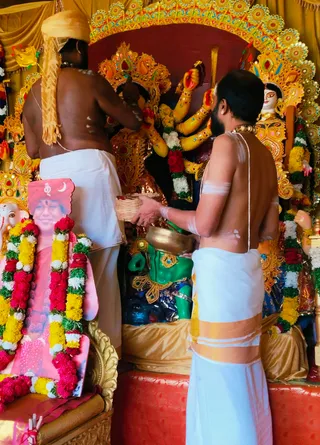 Two bare-chested priests in white dhoti stand before a decorated deity murti of Durga with multiple arms and a golden crown, set against gold and red drapery. The priest at left wears a tall golden turban and works at the figure's raised arms; the priest at right, bearded, holds a brass bowl and a woven basket while wearing a gold-bordered white dhoti and a sacred thread. Marigold and red flower garlands hang around the deity. At left stands a framed pink-backed photograph of a smiling figure in pink robes, garlanded with marigolds, beside a small Ganesha murti. Scene suggests a Durga puja ritual.