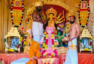A photograph of a Durga Puja shrine at KAILASA Hyderabad with a large gold-and-red deity murti of Durga, multiple arms holding weapons, flanked by a Ganesha murti and a green figure. Two men stand at the shrine: a bare-chested man in white dhoti with a yellow head wrap and rudraksha-style beads performs the ritual holding a sword-like implement, while a second bare-chested man in white dhoti and gold-bordered cloth holds a brass bowl. A pink printed portrait-like image on a stand is garlanded with yellow and red flower garlands, and smaller framed photos on gold side tables are decorated with garlands. Gold-and-multicolor garlands hang on the sides and draped gold cloth forms the backdrop.