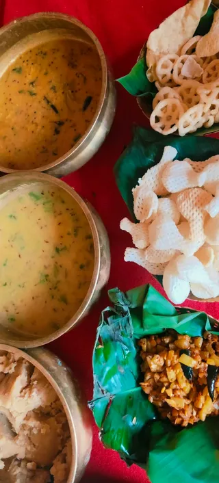 Overhead photograph of a festive vegetarian meal arranged on a red cloth. Three polished metal bowls hold yellow dal, a pale yellow sambar or kadhi with green herb garnish, and a cream-coloured sweet or pudding. Green banana leaves hold crisp snacks, including ring-shaped murukku and white puffed chips, and a portion of spiced potato or vegetable fry wrapped in a leaf. No people, deities or a person are visible in the frame.