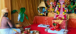 A photograph of a puja set up before a shrine draped in gold cloth. A priest with a shaved head, white dhoti and orange turban sits bare-chested on the left, facing the altar. Brass pots and bowls of kumkum and offerings sit on a red cloth, with a banana plant and a kalasha-style brass pot beside him. On the altar, a large colourful deity murti wearing garlands of marigolds and red flowers stands with a pale face and green hands, and a framed portrait sits behind a smaller garland-draped stand. Lit oil lamps and a small tray of offerings sit in front, with a red-and-gold cloth on a pedestal.