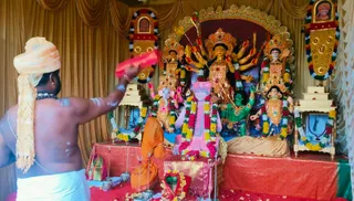 A photograph inside a decorated shrine with gold drapes. In the centre stands a multi-armed goddess murti with a crown and many hands, flanked by smaller deity murtis including an elephant-headed figure and a green-skinned figure. A bare-chested priest with a cream turban and white dhoti stands at left, waving a red cloth over the altar. A pink-draped figure of a child-like deity murti sits in the middle, garlanded with red and yellow flowers. Orange cloth and flower offerings lie on the altar, with oil lamps and framed portraits on the side pillars. Strings of marigolds and red-and-white garlands hang throughout the room.