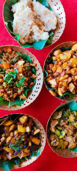 Overhead photograph of five dishes served in perforated paper bowls lined with green leaves on a bright red cloth. Top left holds a mound of pale cooked grain. Middle left has a stir-fry of mixed beans, carrot pieces and green chillies garnished with fresh coriander. Middle right shows diced yellow banana or plantain pieces sauteed with spices. Bottom left contains diced fried potato or plantain with coriander leaves. Bottom right shows a dish of sliced green gourd or vegetable with a dark spiced coating. No people, deities or ritual setting are visible.