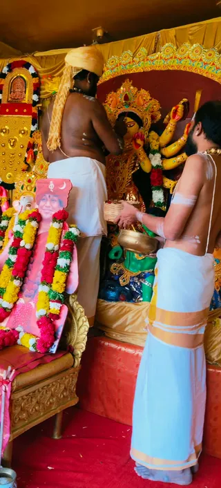 Two bare-chested priests in white dhotis perform puja before a golden-crowned Durga murti with multiple arms holding weapons, draped in red and white garlands. The priest on the left, with a saffron-yellow head cloth and rudraksha mala, stands at the idol; the priest on the right, with a bowl and basket of offerings, holds a brass vessel. On the left a framed portrait with flower garlands shows a smiling figure in pink, next to a golden ornate chair. A gold-framed shrine with a small deity image stands at the far left, and red carpeting covers the floor.